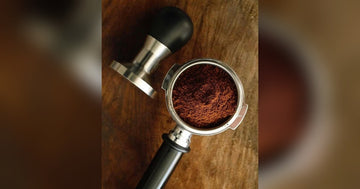 Portafilter with freshly ground coffee and a tamper on a wooden cafe counter